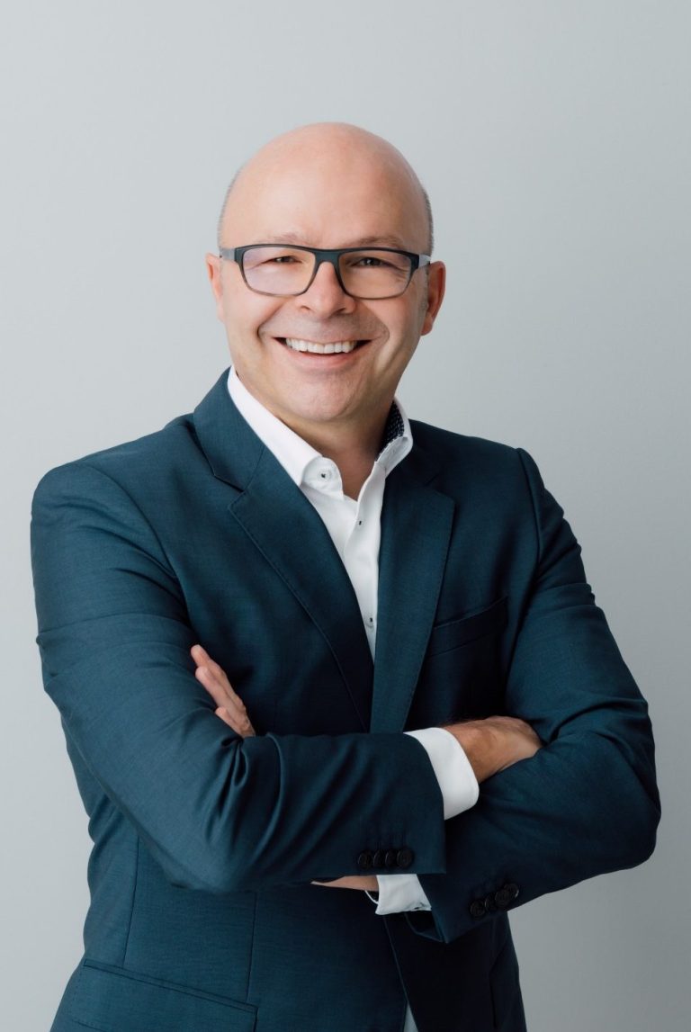 Porträt Simon Hauger Mann mit Glatze und Brille in Anzug, lächelt und steht mit verschränkten Armen.